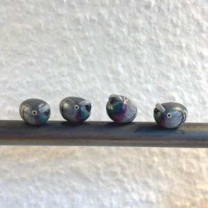 Könnte beinhalten: Vier kleine, abgerundete, handbemalte Taubenfiguren sind horizontal auf einer dunklen Metallstange aufgereiht. Jeder Vogel hat einen grauen Körper mit irisierenden violetten und grünen Halsdetails, einen kleinen weißen Schnabel und ein Auge.