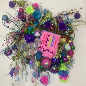 May include: A vibrant Christmas wreath with a pink "Merry Christmas" sign. The wreath is adorned with colorful ornaments, including purple, teal, and green baubles, pom-poms, and a decorative bow. Glittering branches and ribbons add to the festive design.