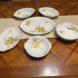 May include: A white porcelain dinnerware set featuring yellow rose floral designs. The set includes serving bowls, oval platters, and a covered soup tureen. Gold trim accents the edges of each piece, adding a touch of elegance to the collection.
