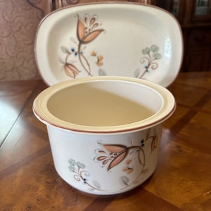 May include: A set of two ceramic dishes with a floral design. The larger dish is oval shaped and the smaller dish is round. Both dishes have a cream background with brown, orange, and green floral accents.