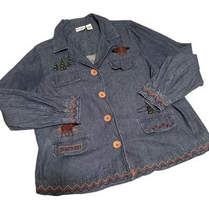 May include: A denim jacket with embroidered forest and animal designs. The jacket features a button-up front, two chest pockets, and two lower pockets. The cuffs and hem are trimmed with a zig-zag pattern. The jacket has a collared neckline and long sleeves.