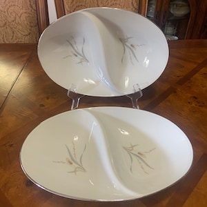 May include: Two white divided serving dishes with a silver rim and a floral design. The dishes are oval shaped and have a divided center.