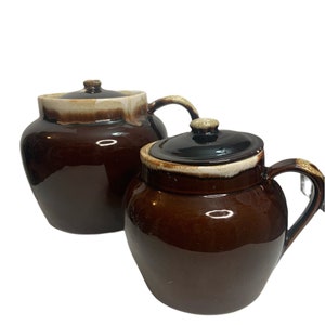 Pode incluir: Duas panelas de cerâmica marrons com tampas. As panelas têm um acabamento brilhante e uma alça. As tampas são marrons escuras com um pequeno botão. As bordas das panelas têm um esmalte marrom claro e branco.