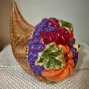 Peut inclure: Une corne d'abondance en céramique avec un motif de panier tressé beige débordant de fruits et légumes sculptés, notamment des raisins violets, des pommes rouges, une citrouille orange vif et des feuilles vertes avec une finition brillante.