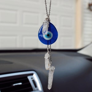 Puede incluir: Un amuleto de ojo turco azul con un centro blanco y una cadena plateada. Dos cristales de cuarzo transparentes cuelgan del amuleto.