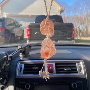 Puede incluir: Un amuleto de coche de cristal de sal rosa del Himalaya colgado del espejo retrovisor de un coche. El cristal est&aacute; atado con una cuerda de macram&eacute; y tiene cuentas de madera en la parte inferior.