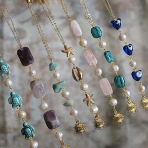 Puede incluir: Collares dorados con varios colgantes, incluyendo piedras preciosas, perlas, estrellas de mar y dijes de tortuga. Los collares presentan una variedad de colores, incluyendo turquesa, rosa y morado. Cada collar tiene una cadena delicada.