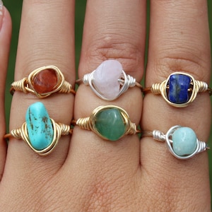 May include: Six rings with different colored gemstones wrapped in gold and silver wire. The gemstones are turquoise, carnelian, rose quartz, green aventurine, lapis lazuli, and aquamarine.