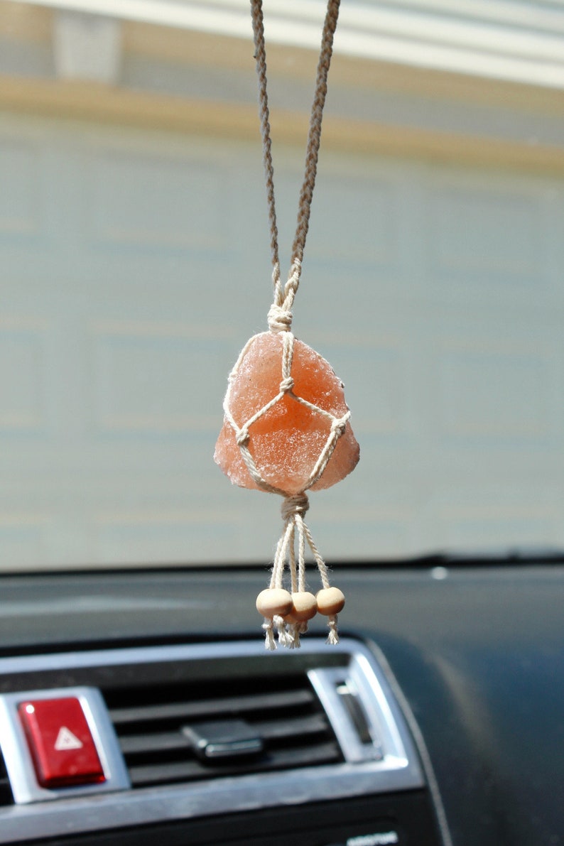 Puede incluir: Una roca de sal rosa del Himalaya est&aacute; suspendida de una macram&eacute; blanca colgante, con tres cuentas de madera en la parte inferior. La colgante est&aacute; unida a un espejo retrovisor en un coche.