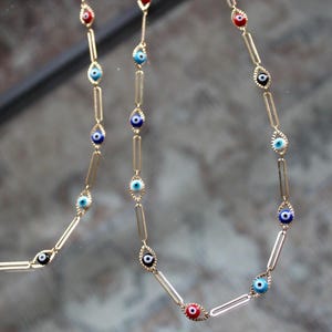 Puede incluir: Dos cadenas de oro con dijes de ojo turco azul, rojo y blanco. Las cadenas están unidas y cuelgan sobre un fondo gris.