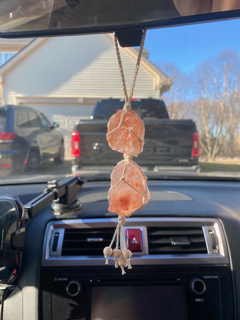 Puede incluir: Un amuleto de coche de cristal de sal rosa del Himalaya colgado del espejo retrovisor de un coche. El cristal est&aacute; atado con una cuerda de macram&eacute; y tiene cuentas de madera en la parte inferior.
