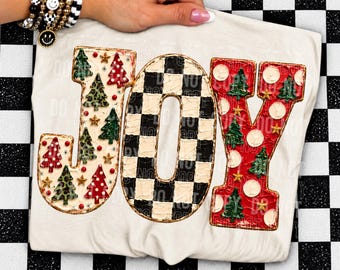 May include: A white t-shirt featuring the word JOY in large, textured letters. The J is cream with green and red trees, the O is a black and white checkerboard pattern, and the Y is red with white polka dots and green trees.