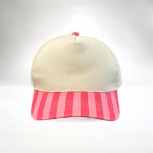 May include: A beige baseball cap with a pink and red striped brim. The cap has a rounded crown and a small pink button on top. The brim features vertical stripes in varying shades of pink and red.