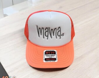 May include: Orange and white trucker hat with the word "mama" in black script across the front. The hat has a mesh back and a curved brim. A small heart is next to the word "mama". The hat has a "World Famous Foam Front" sticker.
