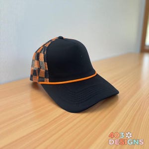 May include: Black baseball cap with an orange and black checkered mesh back panel and a matching orange rope detail along the brim. The cap has a curved brim and a button on top. The text "403 DESIGNS" is visible.