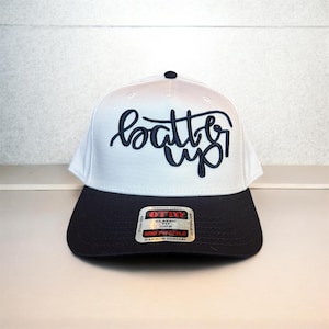 May include: A white and navy blue baseball cap with the words "batter up" in navy blue script. The cap has a curved brim and a button on top. The cap is made of a premium fabric.