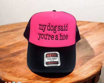 May include: A trucker hat with a pink front panel and a black brim. The text "my dog said you're a hoe" is embroidered on the front. The hat has a mesh back and a snap closure. The brand tag is visible.