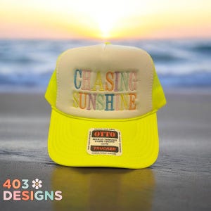 May include: A bright yellow trucker hat with a cream-colored front panel. The text "CHASING SUNSHINE" is embroidered in colorful letters. The hat has a mesh back and a snap closure. The brand label "OTTO" is visible.