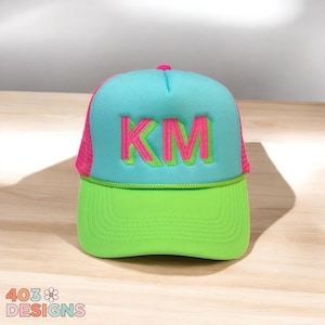 May include: A neon green and turquoise trucker hat with pink mesh sides. The hat features the embroidered letters "KM" in pink. The brim is neon green, and the top is turquoise. The hat is a vibrant accessory.