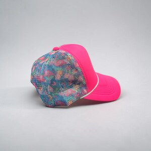 May include: A bright neon pink trucker hat featuring a mesh back panel with a colorful flamingo pattern. The cap includes a solid neon pink front panel, a matching neon pink brim, and a thin white cord detail separating the front and back sections.