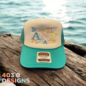 May include: A teal and cream trucker hat with the words "BOATIN AND FLOATIN" embroidered in colorful letters. The hat has a mesh back and a curved brim. The hat is sitting on a wooden surface near the water.