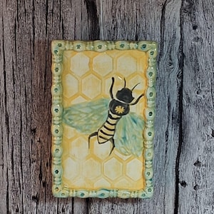 May include: Rectangular ceramic tile with a bee design. The tile features a yellow honeycomb background, a black and yellow bee, and a light green border. The bee has light blue wings. The tile is set against a wooden background.