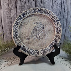 May include: A decorative ceramic plate with a raven design. The plate features a cream-colored background with a detailed raven illustration in the center. The rim is adorned with a repeating spiral pattern in a darker shade. The plate is displayed on a black stand.