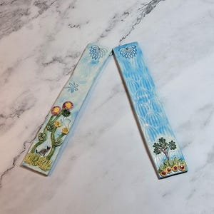May include: Two ceramic bookends with a light blue background. One bookend features a cat, flowers, and a green base. The other has a tree and flowers. Both have a floral design at the top.