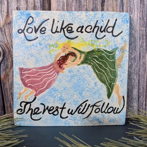 May include: Square ceramic tile with a light blue background and the words "Love like a child, The rest will follow" in black script. The tile features an illustration of two figures embracing, one in a pink dress and the other in a green dress.