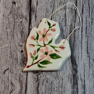 May include: A white ceramic ornament in the shape of the state of Michigan, hand-painted with pink flowers and green leaves. A white string is attached for hanging. The ornament is set against a weathered wood background.