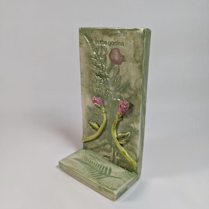 Puede incluir: Un sujetalibros rectangular de cerámica verde claro con un acabado texturizado. Presenta rosas y helechos en relieve, con la inscripción "In the garden of thy heart". Las rosas son rosas y los tallos son amarillos.