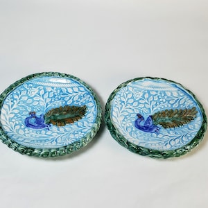 May include: Two ceramic plates with a peacock design. The plates have a light blue background with white floral patterns. Each plate features a blue peacock and a brown feather. The rims are green.