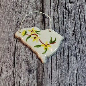 May include: A white ceramic ornament in the shape of the state of South Carolina, with a yellow floral design and a white string for hanging. The ornament is hand-painted with yellow flowers and green leaves.