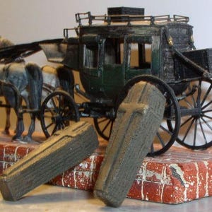 May include: A miniature stagecoach model with a dark green body, black wheels, and a team of gray horses. Two gray coffins with cross details rest on a brick base, creating a unique display.