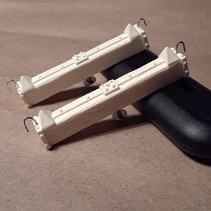 May include: Two cream-colored, rectangular architectural models with decorative details and small metal hooks. The models are resting on a dark, curved object, possibly a remote control, against a neutral background.