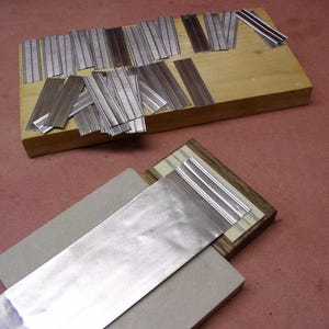 May include: A collection of small, rectangular metal pieces with parallel lines, scattered on a wooden block. Some pieces are arranged on a second wooden structure with a larger metal sheet. The background is a reddish-brown surface.