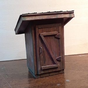 May include: A miniature wooden outhouse model with a weathered, rustic appearance. The structure features a slanted roof, a door secured with a diagonal brace, and a small handle. The wood has a brown and green color.