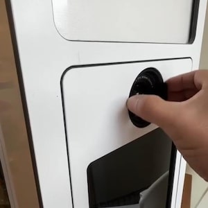 May include: A white photo booth with black trim. A hand is adjusting a black lens on the front panel. The booth has a rectangular window and a lower opening, possibly for a camera or user interaction.