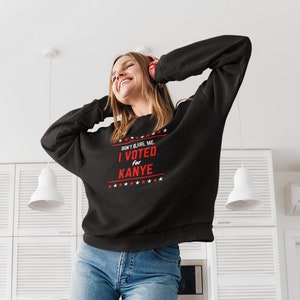 May include: A black sweatshirt with the text "Don't blame me. I voted for Kanye." in red and white. The sweatshirt is being worn by a person standing on a blue couch.