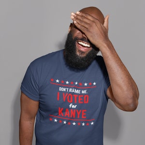 May include: A man wearing a navy blue t-shirt with a red and white graphic that says "Don't blame me. I voted for Kanye." The graphic is surrounded by red and white stars.