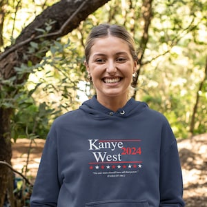 以下が含まれることがあります： 紺色のパーカーを着た人が、赤と白で「Kanye West 2024」と書かれたパーカーを着ています。パーカーには白い紐とカンガルーポケットが付いています。その人は右手に緑色の水筒を持っています。