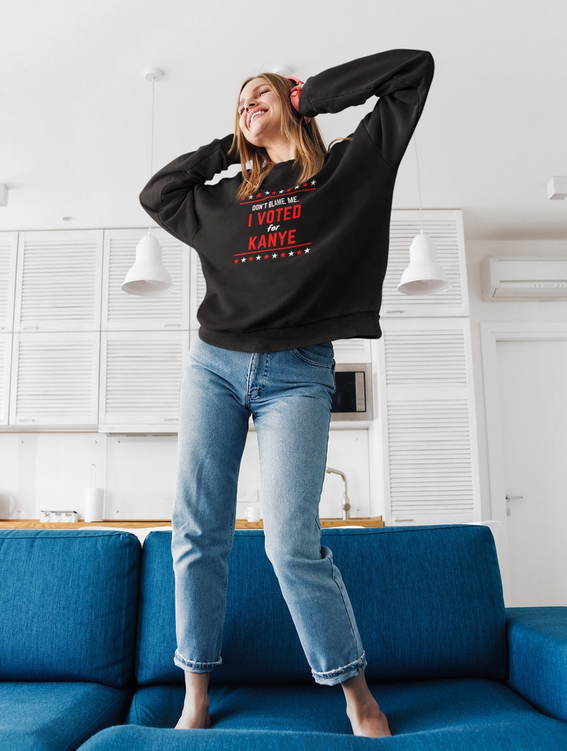 May include: A black sweatshirt with the text "Don't blame me. I voted for Kanye." in red and white. The sweatshirt is being worn by a person standing on a blue couch.