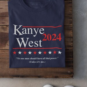 Puede incluir: Una camiseta azul marino con el texto "Kanye West 2024" en blanco y rojo. La camiseta tambi&eacute;n presenta una fila de estrellas blancas y el texto "No one man should have all that power. (Unless it's me.)"