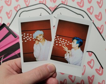 May include: Two Polaroid photos of a person blowing hearts. The person in the left photo has blue hair and is wearing a white shirt. The person in the right photo has blue hair and is wearing a white shirt. Both photos have a red background and white hearts.