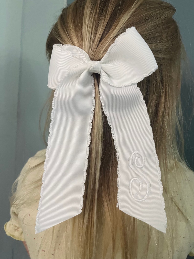 K&ouml;nnte beinhalten: Wei&szlig;es Satin-Haarband mit gewelltem Rand und einem wei&szlig;en gestickten Buchstaben "S" auf einer Seite.