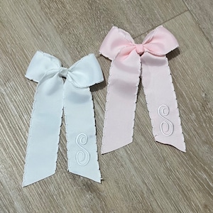 May include: Two hair bows with scalloped edges. One bow is white with a white embroidered letter "S" and the other bow is pink with a white embroidered letter "S".