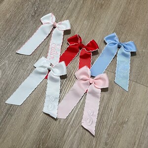 K&ouml;nnte beinhalten: F&uuml;nf Haarb&auml;nder mit gewellten Kanten und bestickten Buchstaben. Die Schleifen sind in Wei&szlig;, Rosa, Rot und Blau. Die Beschriftung umfasst die Namen "Sailor" und "Stella" sowie den Buchstaben "S".