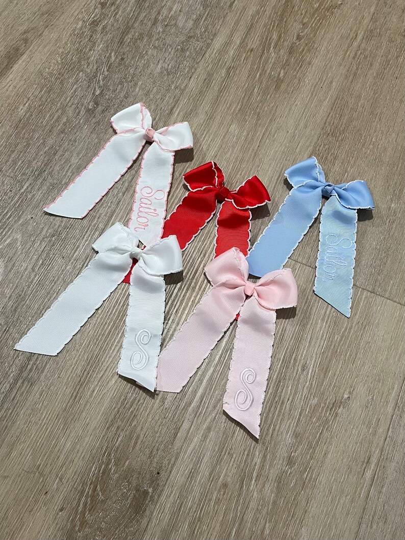 K&ouml;nnte beinhalten: F&uuml;nf Haarb&auml;nder mit gewellten Kanten und bestickten Buchstaben. Die Schleifen sind in Wei&szlig;, Rosa, Rot und Blau. Die Beschriftung umfasst die Namen "Sailor" und "Stella" sowie den Buchstaben "S".