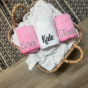 May include: A woven basket holds folded white and pink towels. The pink towels are embroidered with the names "Lina" and "Tessa" in silver thread. A white towel embroidered with "Kate" is also in the basket. The basket sits on a wood floor.