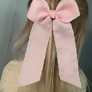 K&ouml;nnte beinhalten: Ein rosa Haarb&auml;ndchen mit einem wei&szlig;en gewellten Rand und einem wei&szlig;en gestickten Buchstaben "S".